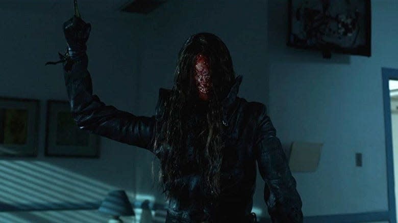 A person with a disfigured face stands in a dark room, holding up a pointed object, dressed in black, long hair covering parts of their face