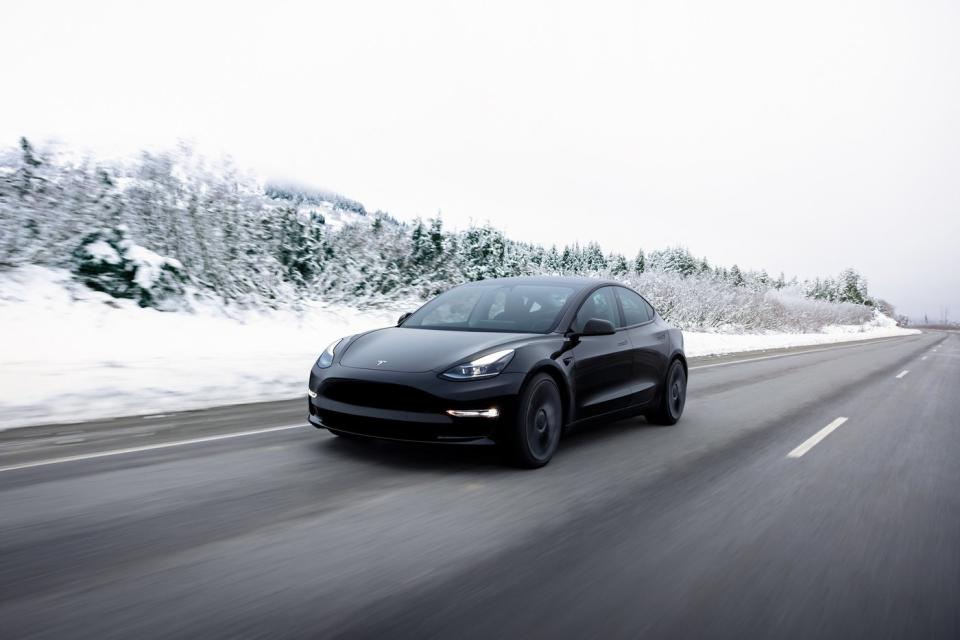 A black Tesla car drives on an open road in the snow.