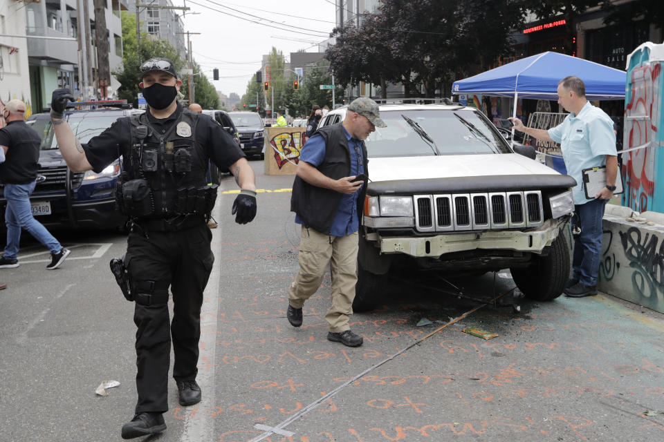 A Seattle police officer asks people to move aside to allow police vehicles through as investigators, right, look over a car involved in a shooting Monday, June 29, 2020, in Seattle, where streets are blocked off in what has been named the Capitol Hill Occupied Protest zone. One man was killed and another wounded early Monday in the protest zone, the second deadly shooting in the area. Police said the shooting happened before dawn in the city's Capitol Hill neighborhood, near downtown. (AP Photo/Elaine Thompson)