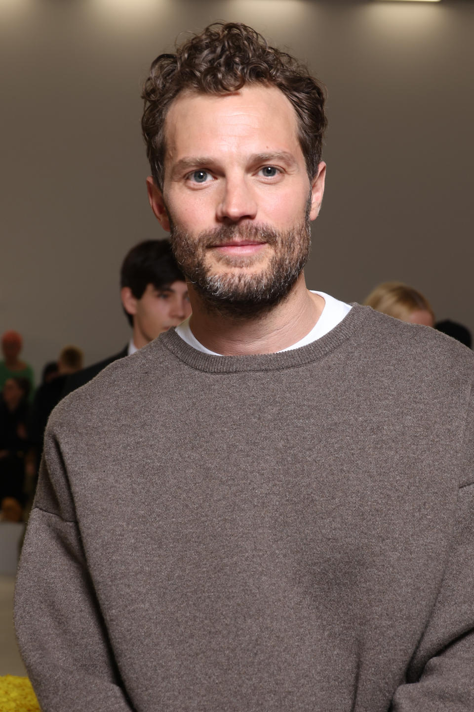 Jamie Dornan wearing a casual sweater, standing in a crowded indoor event space