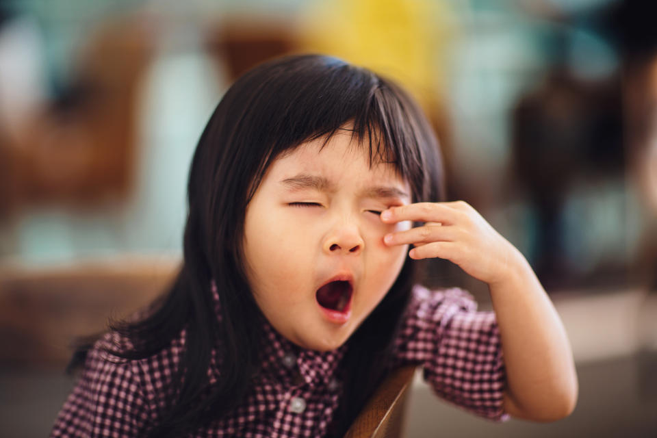 Sleepy toddler girl resting her arm in wooden chair while yawning & rubbing her eyes in a cafe.