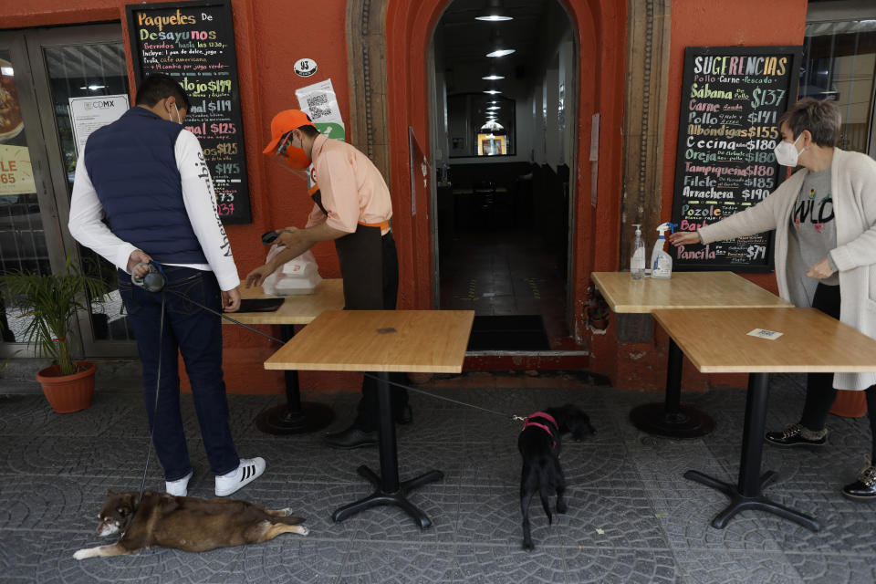 Waiter Luis Angel Garcia Gonzalez delivers a takeout order to a customer as General Director Mireya Ruiz reaches for hand sanitizer, outside the Condesa location of La Casa de la Yeya restaurant, which has already had to shutter three of its 10 locations amid the COVID-19 pandemic with two more under threat, in Mexico City, Sunday, Jan. 10, 2021. Worried about their ability to survive, many restaurants in the capital and adjacent Mexico State are banning together in a campaign dubbed "Abrir o Morir," Spanish for "Open or Die," and plan to open their doors to diners again on Monday in defiance of ordinances limiting restaurants to takeout service while the Mexico Valley remains under red alert with hospitals nearing full capacity. (AP Photo/Rebecca Blackwell)