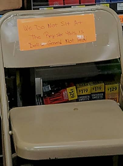 Handwritten sign on a chair stating 'We Do Not Sit At The Register This Is Dollar General Not Aldi' indicating seating not allowed