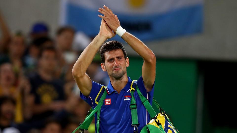 novak djokovic claps his has over his head, he carries two tennis bags and wears a blue polo shirt, his face is red and scrunched from crying