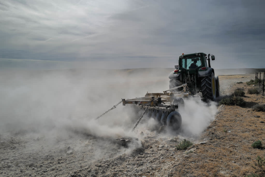 A tractor pulling a plow kicks up clouds of dust in a barren field.