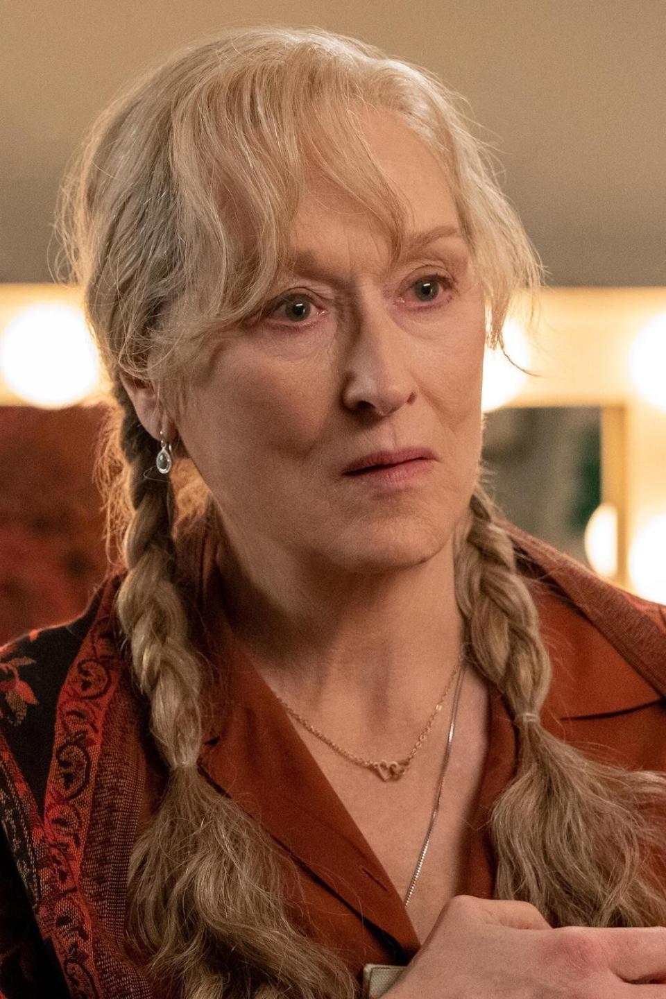 Meryl Streep, in a patterned shawl and braids, holds an envelope and engages in a discussion with another person in a dressing room with a lit vanity mirror