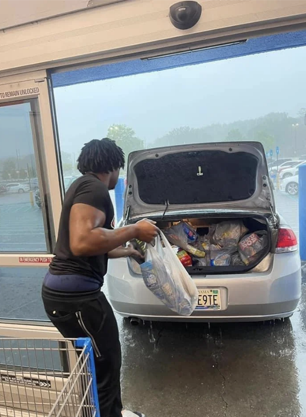 A person loads groceries into the trunk of a car in a parking lot during rain