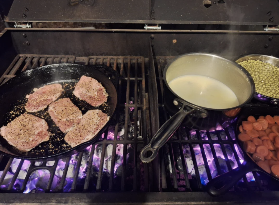 Food cooking on a grill with seasoned meat in a skillet, a pot of creamy sauce, a pot of peas, and sausages in another skillet