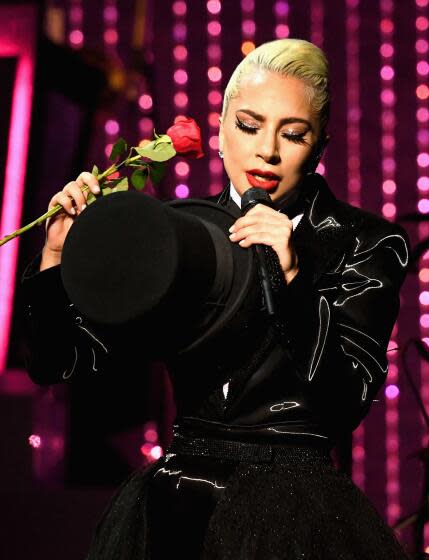 Lady Gaga sings into a microphone with her eyes closed and is wearing a black shiny suit holding a top hat and rose.