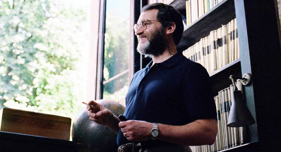 Jon Stone stands near a window, wearing glasses and a dark shirt, gesturing with his hand. He is surrounded by shelves filled with books