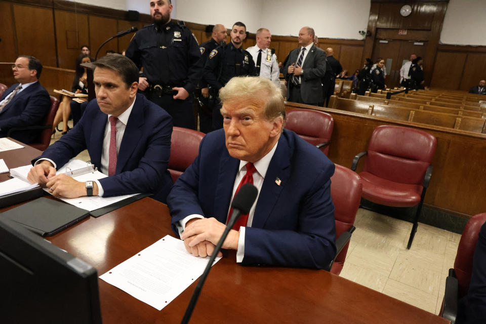 Individual seated at a table with papers in a courtroom setting, surrounded by security personnel