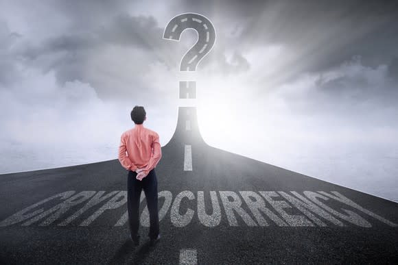 A man stands on a highway which rises up into the sky to end in a large question mark. The pavement is marked with the text Cryptocurrency.