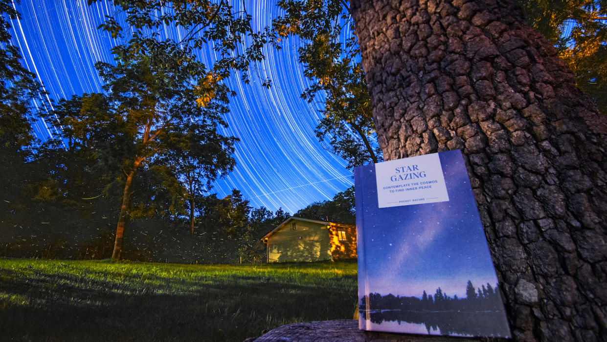  A book leans against a tree against the background of a house in the distance and a dark grassy yard. The night sky is filled with the streaks of stars, traced through the sky through long exposure photos. 