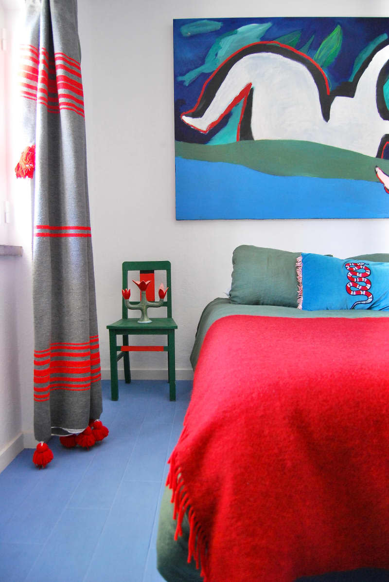 Artwork hung above bed with red blanket in white bedroom with gray curtains and blue floor.