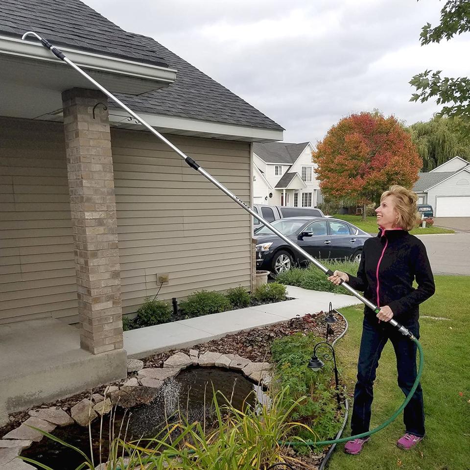 Woman in tracksuit using a telescoping pole to clean a home's rain gutters without a ladder.