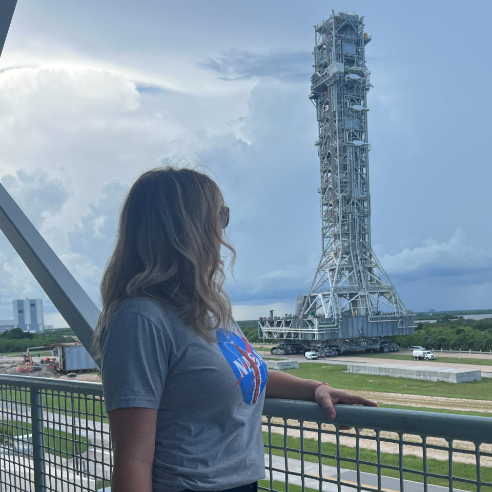 a woman stands, looking at a large metal tower. A square building is seen small in the background.