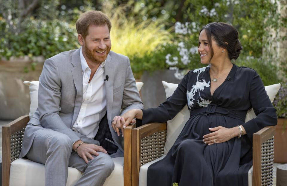 Prince Harry in a casual suit and Meghan, Duchess of Sussex, in a black dress, hold hands