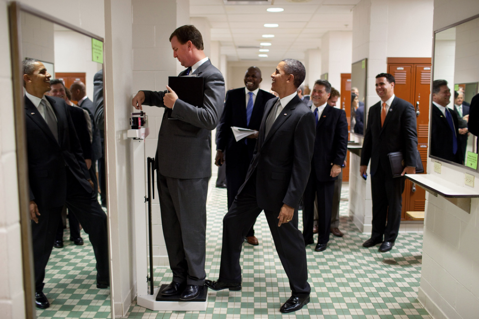 El presidente Obama bromea con Marvin Nicholson, miembro de su equipo, poniendo la puntera del zapato en la balanza mientras este se pesa, sin que lo note, cuandos el séquito presidencial pasaba a través de los vestidores del equipo de Voleibol de la Universidad de Texas en Austin. (Pete Souza/The White House)