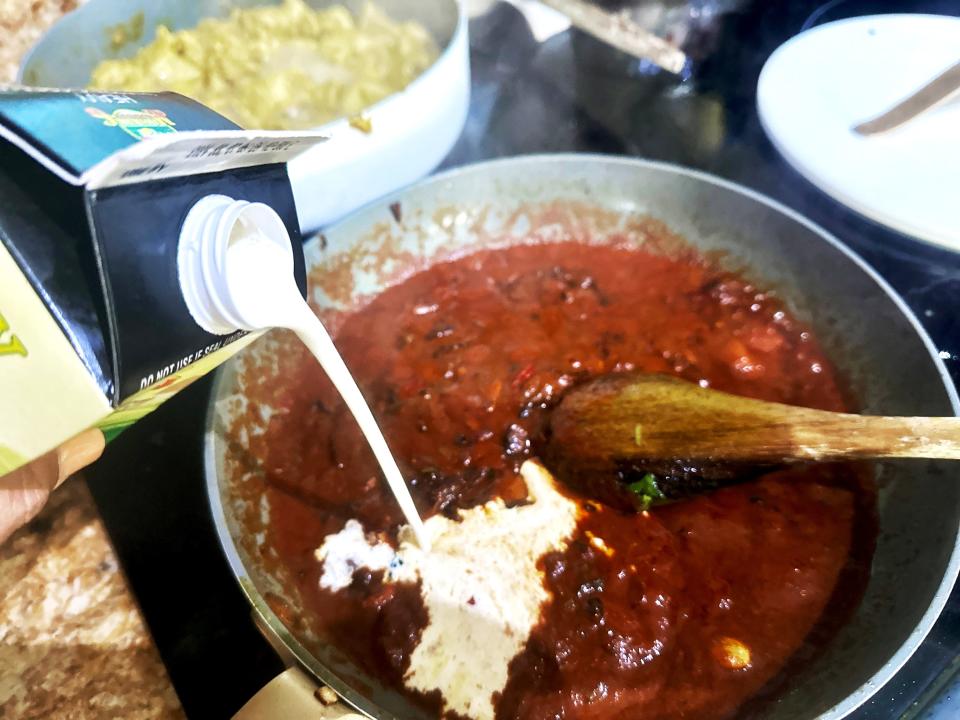 A carton of heavy whipping cream being poured into a pan of chiles and tomato sauce. A wooden spoon sticks out of the pan