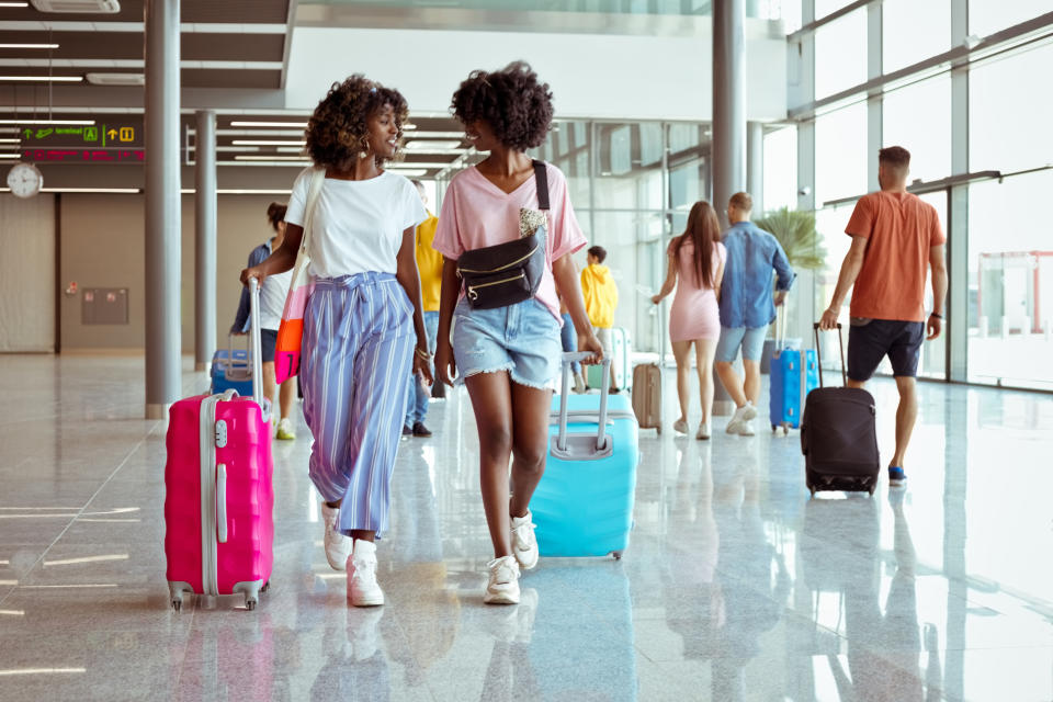 Two women walking in an airport with suitcases, each dressed in casual travel outfits including shorts, skirts, and comfortable tops. Other travelers are in the background