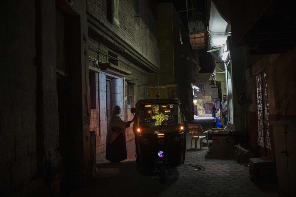 In this Nov. 19, 2019 photo, a woman talks to a tuk-tuk driver in an alleyway of a slum in Cairo, Egypt. Motorized rickshaws known as tuk-tuks have ruled the streets of Cairo’s slums for the past two decades hauling millions of Egyptians home every day. Now the government is taking its most ambitious stand yet against the polluting three-wheeled vehicles: to modernize the neglected transport system, it plans to replace tuk-tuks with clean-running minivans. (AP Photo/Nariman El-Mofty)