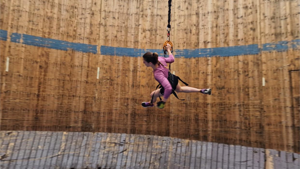  Picture of a female runner wearing a long-sleeve pink top, black shorts and purple and black trainers. She is running along a "Wall of Death" which appears to be made of wood. There is a blue stripe of paint running across the circumference of the wall. She is also holding on to bungee cords and is wearing a harness. 