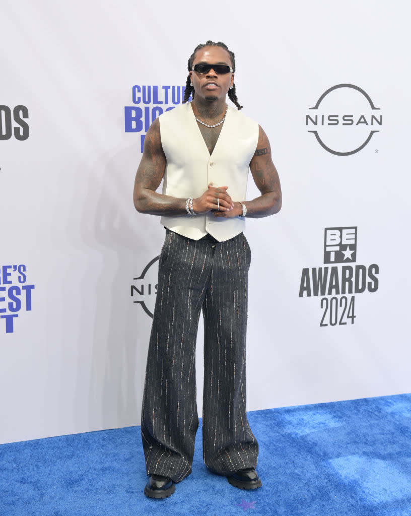 Gunna in a sleeveless vest, wide-legged pants, and sunglasses poses on the red carpet at an event. The backdrop features "BET Awards 2024" and sponsor logos