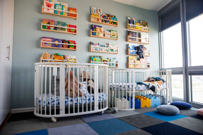 children's room in gray blue with 2 white cribs. shelves of colorful books on the wall above. Blue rug with square pattern. Oversize legos under one of the cribs