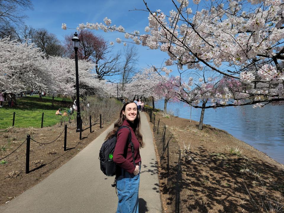 Amanda wears a small backpack and smiles with a park path behind her and cherry blossoms overhead.