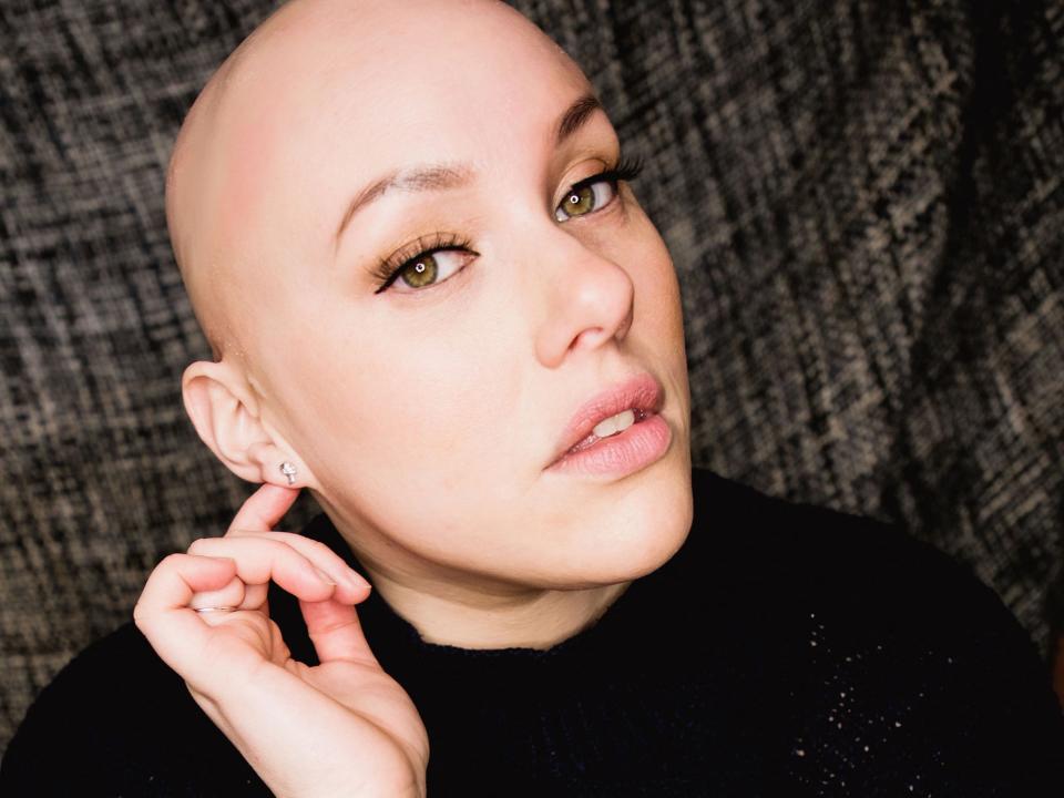 Laura Mathias sits, looking serious with her right hand posed by her ear and the other resting in her lap. She is bald with hazel eyes and wears a black turtleneck.