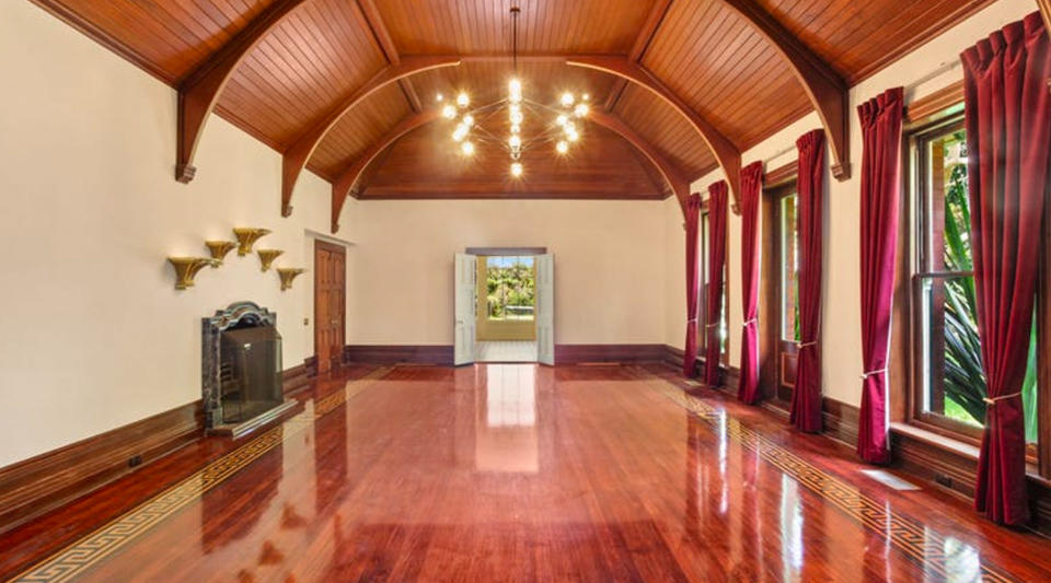 A large room with a high ceiling, polished timber floors, a fireplace and red velvet curtains.