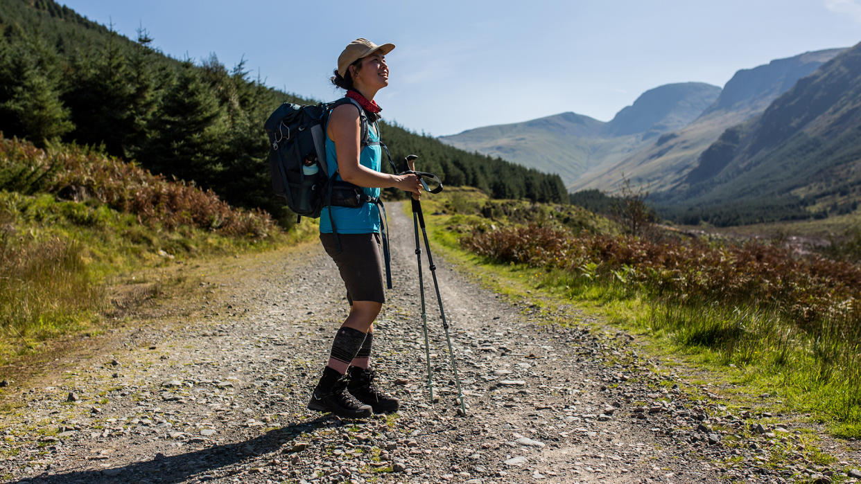  A woman stands on a rocky hill path, holding trekking poles and wearing Salewa Ortles Ascent Mid Gore-Tex Boots. 