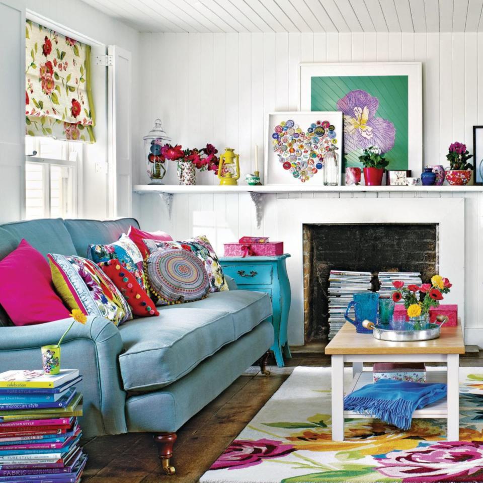 White living room, blue sofa with bright multicoloured cushions, bold floral rug, wooden coffee table, white sofa, stacked books used as table, stacked magazines in fireplace, wooden floorboards, 3D heart art on shelf, potted house plants.