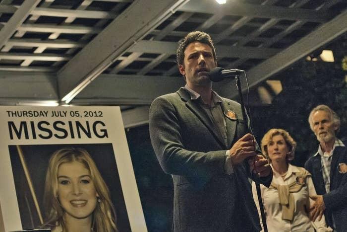 In a scene from the movie, Nick is standing in front of a microphone with a "Missing" poster of his wife