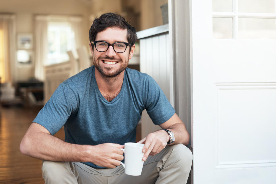 Smiling man with short dark hair and glasses, wearing a casual shirt and khaki pants, seated inside near an open door, holding a coffee mug