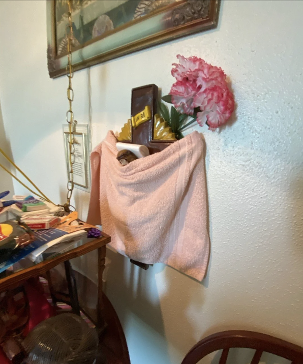 A cluttered interior scene with a towel hanging on a wall, a picture frame, artificial pink flowers, and various household items on a nearby table