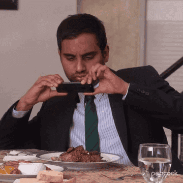 Aziz Ansari in a suit photographing his meal with a phone at a dinner table