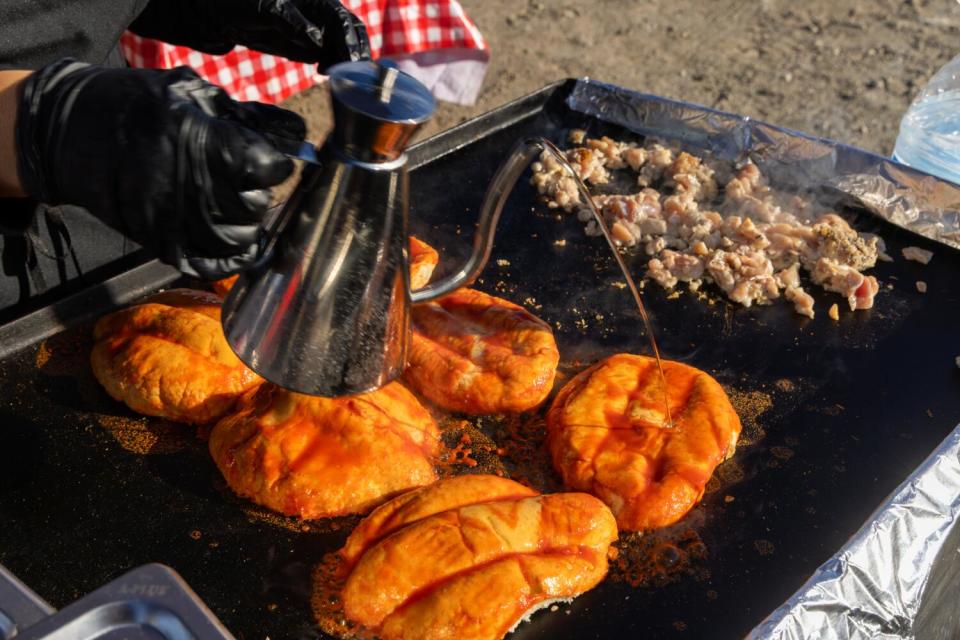 A hand holding a container pours a liquid on meat on a grill outdoors.