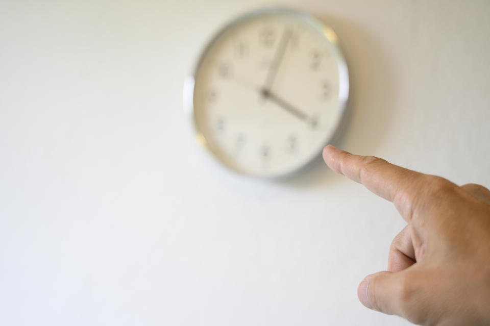 A person's index finger pointing towards a wall-mounted clock indicating a time around 10:10