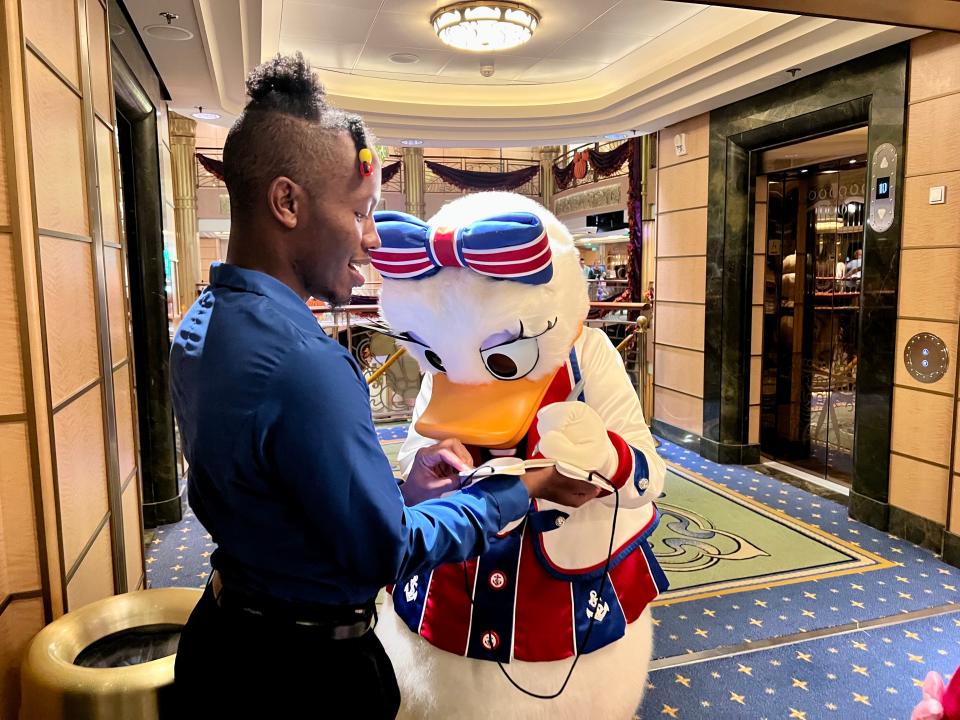 Daisy Duck signs an autograph on a cruise ship, wearing a red, white, and blue bow and shirt. 