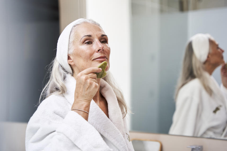 Mature woman doing gua sha massage with gua sha tool in mirror