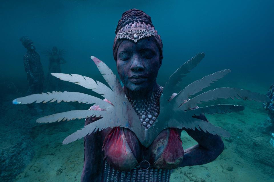 An underwater sculpture of a woman with her eyes closed, wearing an ornate headpiece, feathers, and an outfit splattered in red.