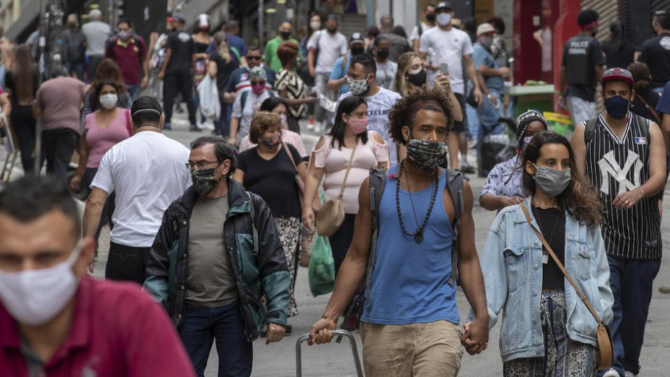 Photo shows a crowd of people, many wearing masks, walking along a busy street in Brazil.