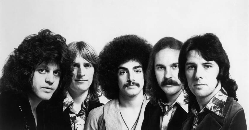 A black and white photo of Journey's founding members in '70s style attire and hairdos looking forward