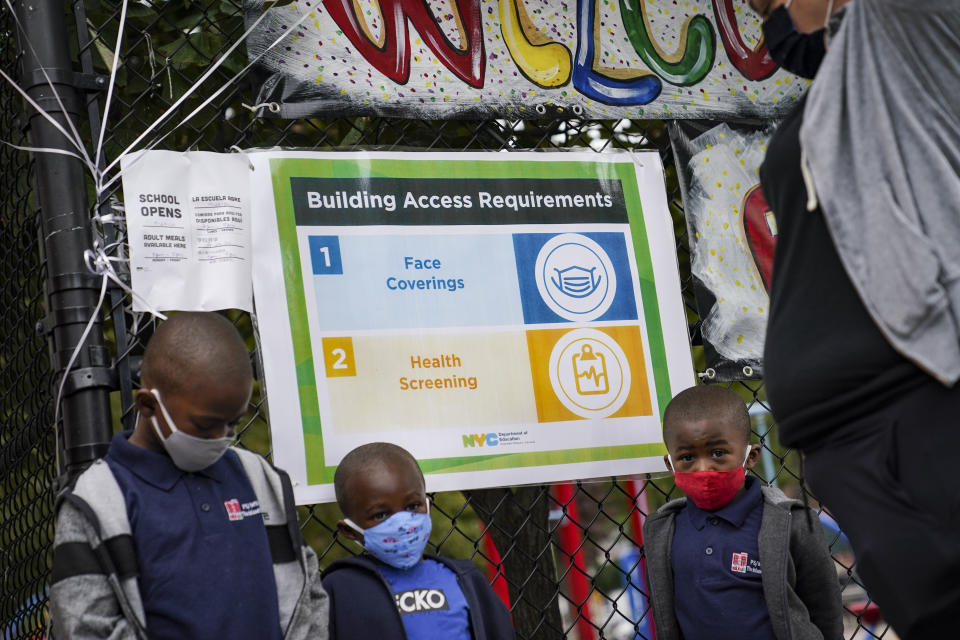 A sign displaying protocols for combating COVID-19 hangs beside students arriving for in-person classes outside Public School 188 The Island School, Tuesday, Sept. 29, 2020, in the Manhattan borough of New York. Hundreds of thousands of elementary school students are heading back to classrooms starting Tuesday as New York City enters a high-stakes phase of resuming in-person learning during the coronavirus pandemic. (AP Photo/John Minchillo)