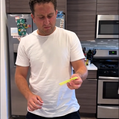 Man in a kitchen looking puzzled at a yellow wrapped item next to a sandwich