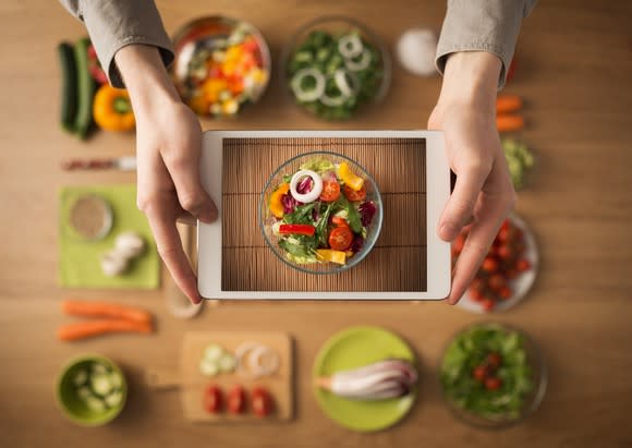 Culinary ingredients spread around wood table, with hands holding a tablet showing the image of the completed dish, a colorful salad.