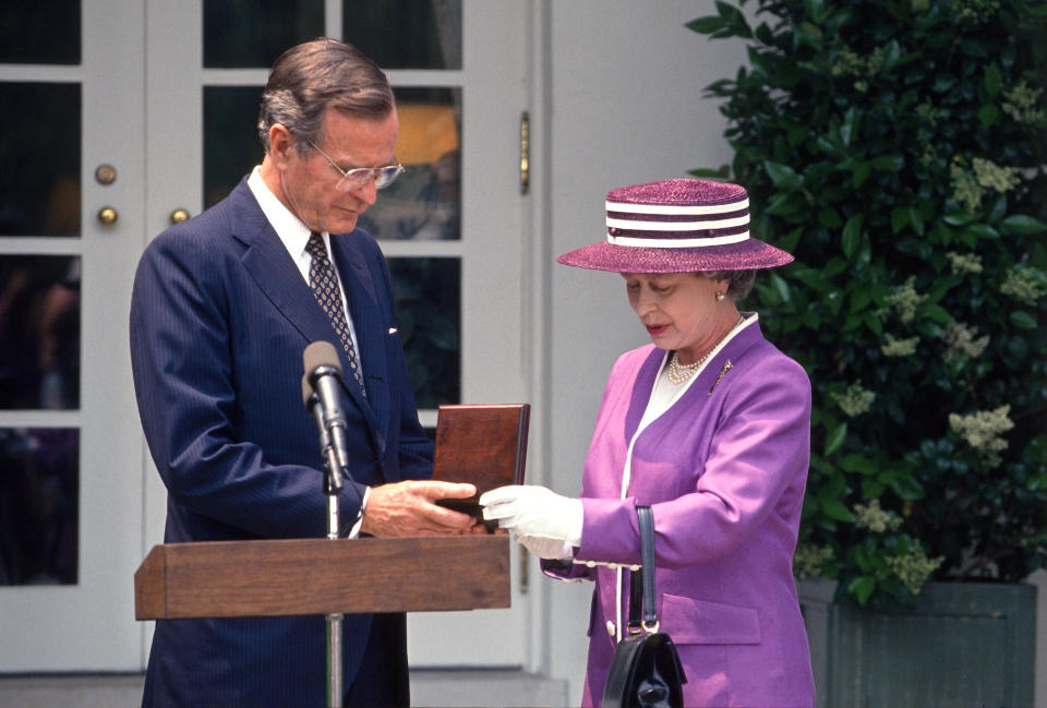 Queen Elizabeth II, in purple outfit and purple striped hat, presents President George H.W. Bush a case containing a medal in front of a microphone.