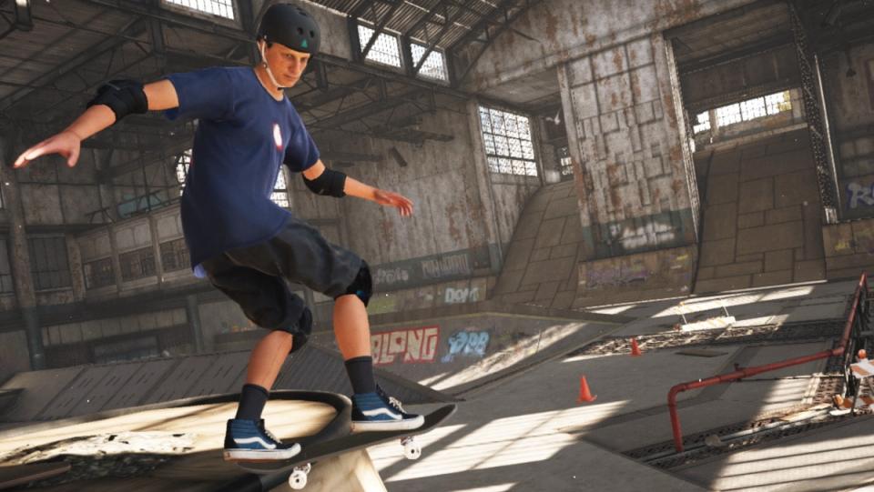 The video game version of Tony Hawk wears a blue shirt, shorts, and a helmet, and balances on his skateboard in the game's iconic first stage. Behind him is a huge skate arena full of quarter pipes, ramps, and rails.