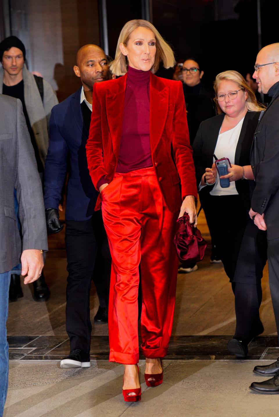 Dion in a velvet red blazer, burgundy turtleneck, bright red satin trousers, red velvet platform heels, and a burgundy satin purse.